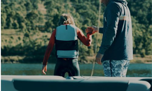 Male adult holding the hand of a child wearing a life jacket, stood near a river bank.
